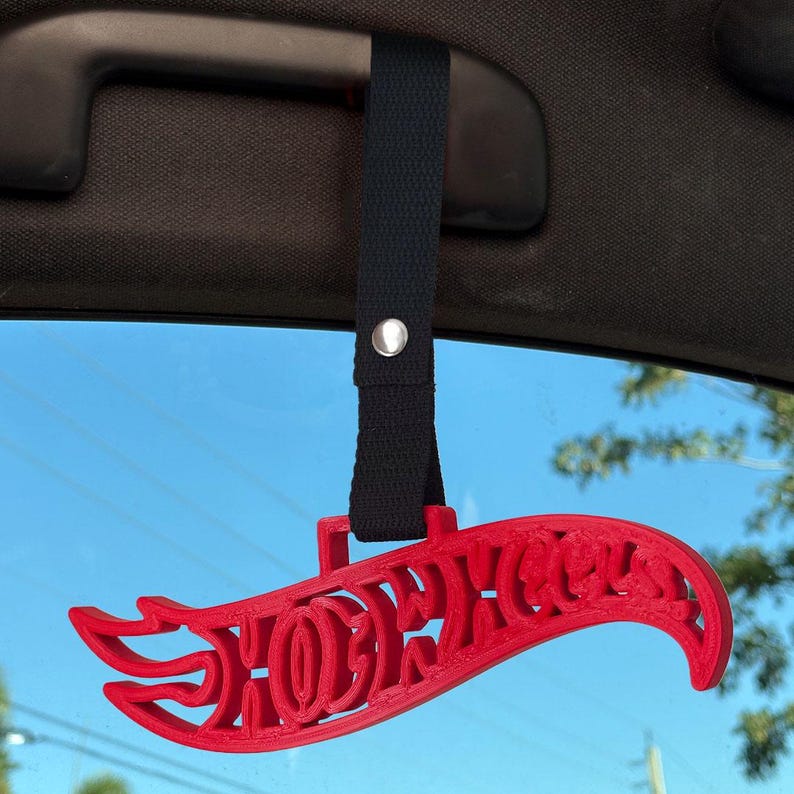 May include: A red 3D-printed Hot Wheels logo hanging from a black fabric strap with a silver metal snap button. The strap is looped around a dark gray car interior grab handle, displaying the iconic flame-shaped brand lettering.