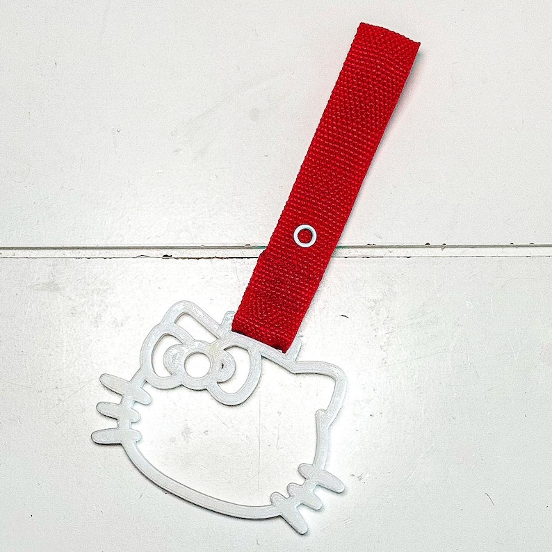 May include: A white Hello Kitty-shaped tag with a red fabric strap. The tag has a cut-out design of the iconic character's face, including her bow and whiskers. The red strap is attached with a white grommet.