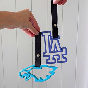 May include: Two blue sports team logo keychains with black straps. One keychain features the letters "LA" in a stylized font, and the other is a blue jaguar logo. The keychains are held up against a white background.