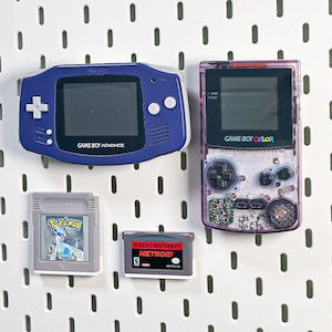 Peut inclure: Une collection de consoles de jeux portables rétro et de cartouches. Comprend une Game Boy Advance bleue, une Game Boy Color violette et deux cartouches de jeu : "Pokemon" et "Metroid". Les consoles et cartouches sont présentées sur fond blanc.