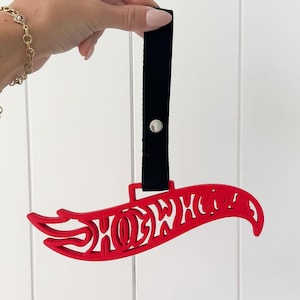 Puede incluir: Un adorno rojo con forma de logotipo de Hot Wheels con una correa negra y un botón plateado. El adorno está sostenido por una mano sobre un fondo blanco. El logotipo de Hot Wheels es una llama estilizada con el nombre de la marca.