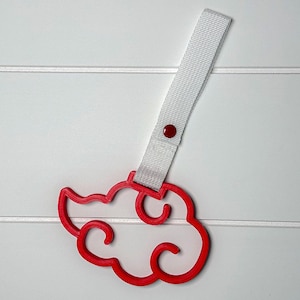 May include: A red cloud-shaped accessory with a white strap and a red button. The cloud design has a curved outline. The strap is attached to the cloud with a red button. The background is white.