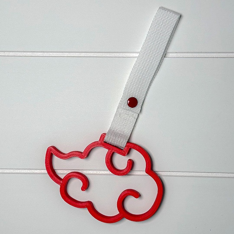 May include: A red cloud-shaped accessory with a white strap and a red button. The cloud design has a curved outline. The strap is attached to the cloud with a red button. The background is white.