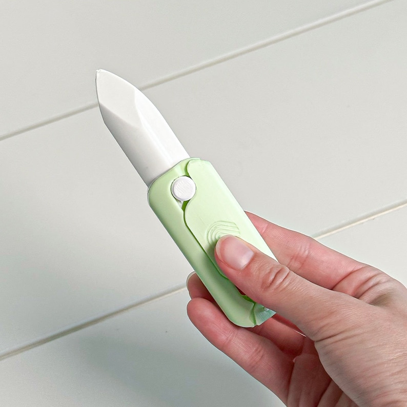 May include: A light green folding knife with a white blade. The knife is held in a hand and has a white button to open and close the blade. The handle is a light green color.