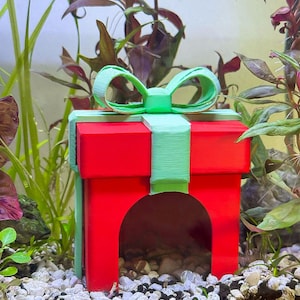 May include: A red and green aquarium decoration shaped like a gift box. The box has a green bow and ribbon, with an arched opening. The decoration is surrounded by aquatic plants and small rocks, creating a natural underwater scene.