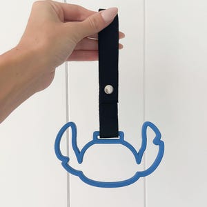 May include: Blue plastic car seat handle with a black fabric strap and a silver snap. The handle is in the shape of a cartoon character. The strap is held by a hand.