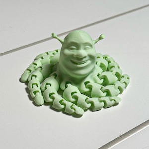 May include: A light green 3D-printed sculpture of a smiling ogre's face with small antennae, surrounded by a ring of segmented tentacles. The sculpture is on a white surface.