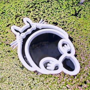 May include: White 3D printed Totoro character cutout floating in a pond of green and pink water plants.