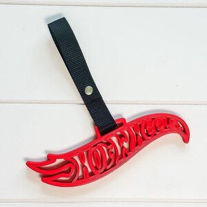 May include: A red plastic Hot Wheels logo keychain featuring a black nylon strap attached with a silver metal rivet. The iconic flame-shaped text is cut out in a bold, stylized font, providing a lightweight and durable accessory.
