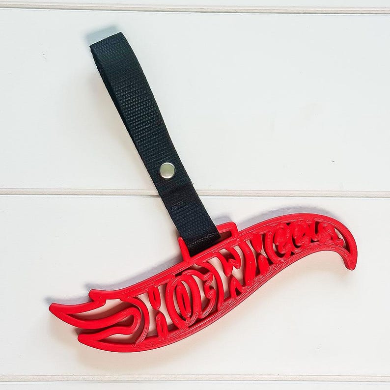 May include: A red plastic Hot Wheels logo keychain featuring a black nylon strap attached with a silver metal rivet. The iconic flame-shaped text is cut out in a bold, stylized font, providing a lightweight and durable accessory.