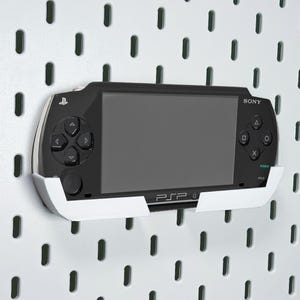 Może przedstawiać: Czarna konsola do gier przenośna Sony PSP zamontowana na białym wsporniku, na tle białej płyty perforowanej. Konsola posiada duży ekran, pad kierunkowy i przyciski akcji. Wspornik został zaprojektowany tak, aby bezpiecznie trzymać konsolę.