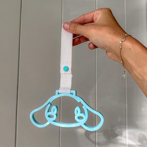 May include: A light blue, cartoon-style handle with a white strap and a teal button. The handle is held up by a hand. The handle has a dog-like face design.