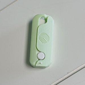 May include: A light green, plastic key holder with a rounded top and a white button. The key holder has a curved design with a circular logo on the front.