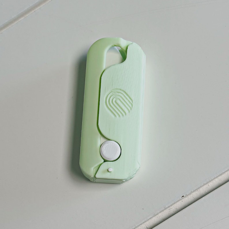 May include: A light green, plastic key holder with a rounded top and a white button. The key holder has a curved design with a circular logo on the front.