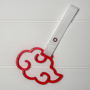 May include: A red cloud-shaped charm with a white outline and a white strap. The charm has a circular hole for attachment. The design is a stylized cloud, likely for a keychain or accessory.