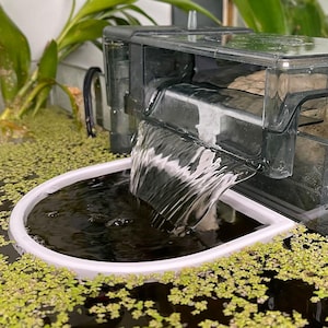 May include: A clear, rectangular aquarium filter with water cascading into a white, oval-shaped basin. The water is dark, and the basin is surrounded by small green plants. The filter is positioned above the basin, creating a waterfall effect.