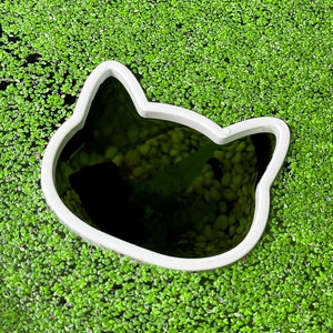 May include: A white ceramic cat-shaped cookie cutter sits on a bed of green moss.