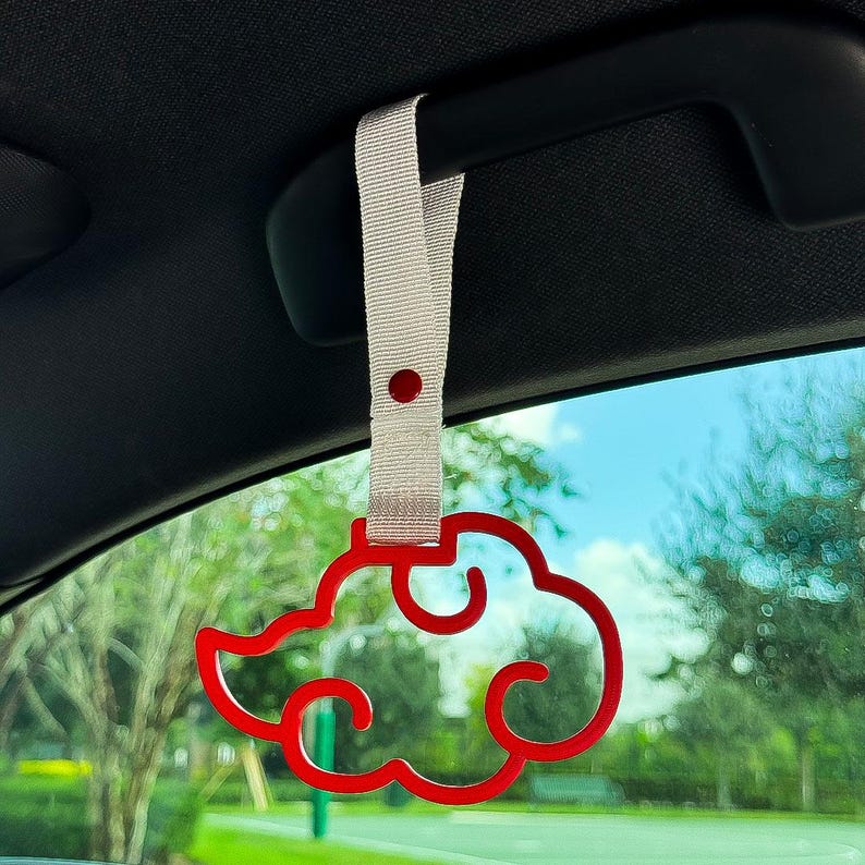 May include: A red cloud-shaped car charm hanging from a light gray strap. The charm is suspended from a car's interior handle, with a scenic outdoor view visible through the window.