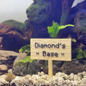 May include: A beige sign with black text that reads "Diamond's Base" is placed in a fish tank with gravel and green plants.