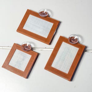 K&ouml;nnte beinhalten: Drei braune, rechteckige Rahmen mit wei&szlig;en Stoffeins&auml;tzen und transparenten Saugn&auml;pfen. Die Rahmen sind auf einer wei&szlig;en Oberfl&auml;che angeordnet und zeigen ein schlichtes, klares Design. Die Rahmen sind wahrscheinlich f&uuml;r Fotos oder Kunstwerke.