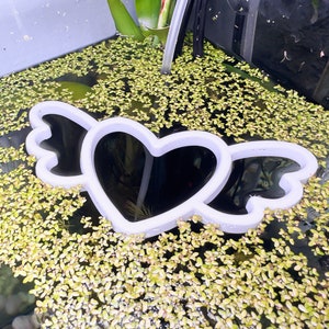 May include: White plastic heart-shaped ornament with wings, floating in a fish tank filled with green aquatic plants.