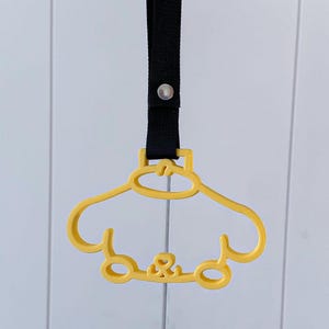 May include: A yellow plastic car-shaped grab handle featuring a cutout design and a black fabric strap attached with a silver metal rivet. The handle has a rounded top section and a lower base with two circular wheel-like shapes at the bottom.