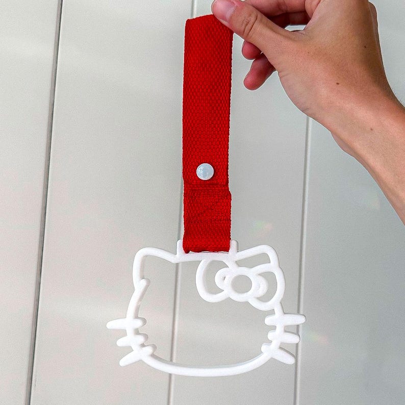 May include: A white Hello Kitty-shaped handle with a red strap. The strap is attached to the handle with a white button. The handle is held up by a hand.
