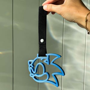 May include: A blue, 3D-printed Sonic the Hedgehog character keychain. The keychain is attached to a black fabric strap with a white button. The character's outline is a vibrant blue, and the design is a recognizable silhouette.