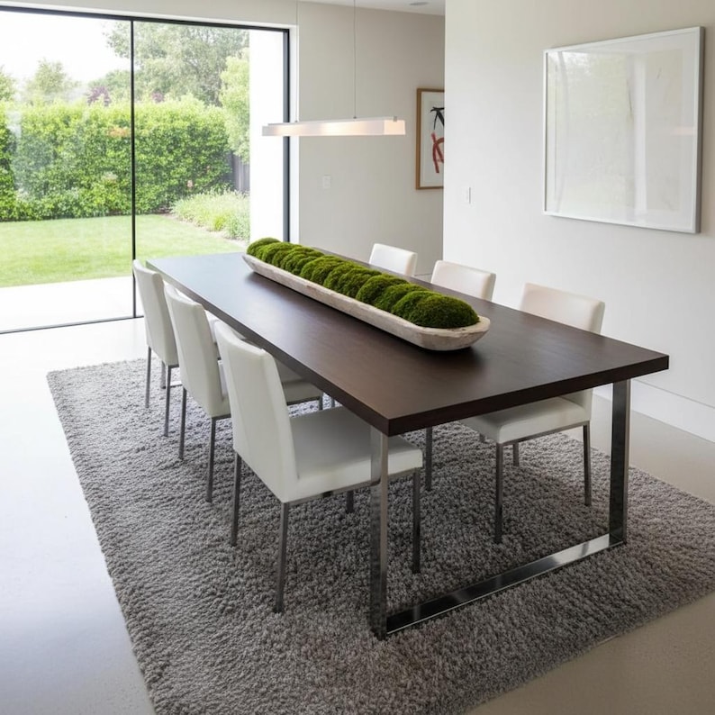 May include: A long, narrow, light-colored wooden dough bowl filled with vibrant green moss, placed as a centerpiece on a dark brown rectangular dining table with metal legs, surrounded by six white chairs on a gray textured area rug.