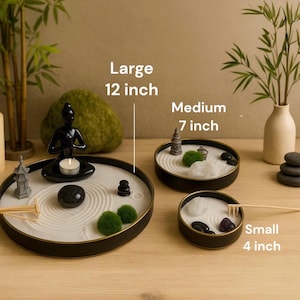 May include: A black circular tray with a sand garden inside. The tray has a small rake, a grey stone, two green bushes, a small black statue of a person sitting in a meditative pose, and three black stones stacked on top of each other.