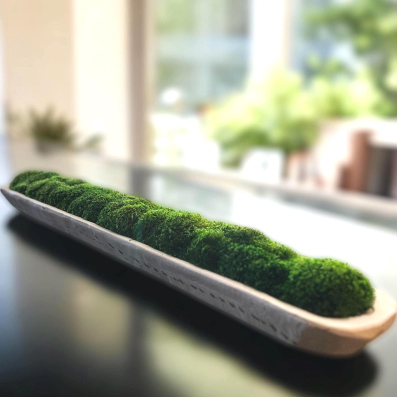 May include: A long, narrow, light-colored wooden dough bowl filled with a continuous row of vibrant green preserved moss. The bowl has a smooth, elongated shape with slightly curved edges, contrasting with the textured, dense green moss.