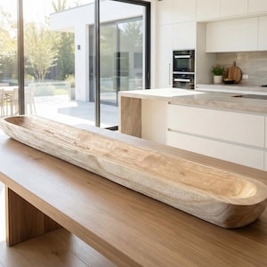 Puede incluir: Un cuenco largo de madera de color claro descansa sobre una mesa de madera. El cuenco tiene un acabado liso y natural y una forma ovalada alargada. La mesa también es de madera y tiene un color marrón claro. El fondo muestra una cocina moderna.