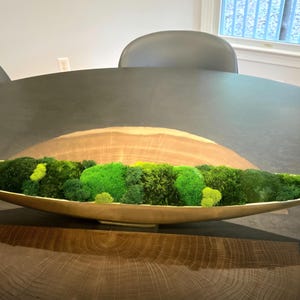 May include: A long, boat-shaped gold metal bowl filled with various shades of preserved green moss. The bowl features a smooth, reflective metallic interior and exterior, contrasting with the textured, organic appearance of the vibrant green moss.