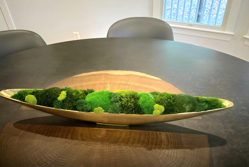 May include: A long, boat-shaped gold metal bowl filled with various shades of preserved green moss. The bowl features a smooth, reflective metallic interior and exterior, contrasting with the textured, organic appearance of the vibrant green moss.