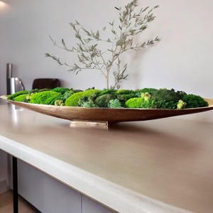 以下が含まれることがあります： さまざまな緑色の苔と小さな植物で満たされた、長い金色の金属製のボウルが特徴の装飾的なセンターピース。細い木の枝が中央から立ち上がり、アレンジメントに高さと自然な質感を与えています。ボウルは明るい色の表面に置かれています。