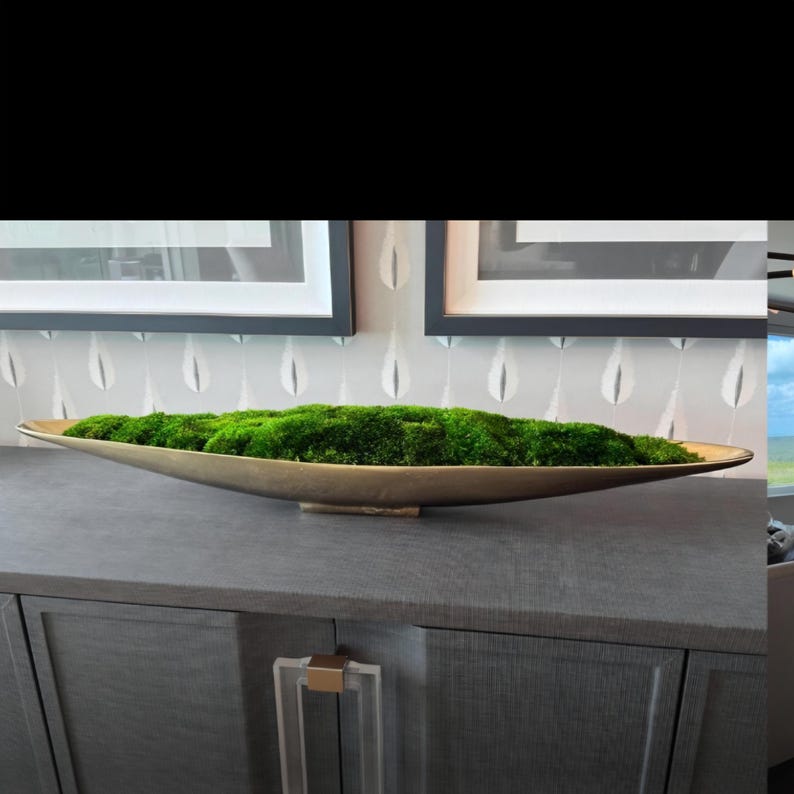May include: A long, shallow, metallic gold-toned oval bowl filled with a dense layer of vibrant green preserved moss. The bowl features a smooth, curved exterior and sits horizontally, showcasing a naturalistic texture within the vessel.