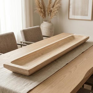 May include: A long, rectangular wooden bowl with a shallow, trough-like interior. The bowl is made of light-coloured wood and sits on a table with a neutral-coloured runner. The overall aesthetic is minimalist and natural, suitable for home decor.