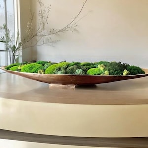 May include: A long, shallow, oval-shaped bowl with a metallic bronze finish filled with a dense arrangement of preserved green moss in various shades, including bright lime, deep forest green, and pale yellow-green textures.