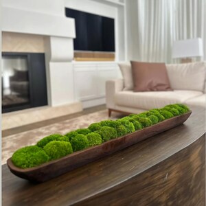 May include: A long, narrow, dark brown wooden dough bowl filled with a row of vibrant green preserved moss balls. The bowl has a smooth, curved interior and a rustic, elongated shape, showcasing the natural wood grain and texture throughout.