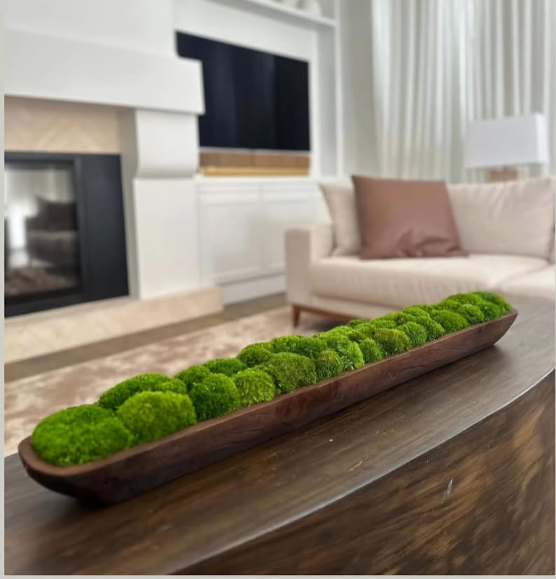 May include: A long, narrow, dark brown wooden dough bowl filled with a row of vibrant green preserved moss balls. The bowl has a smooth, curved interior and a rustic, elongated shape, showcasing the natural wood grain and texture throughout.