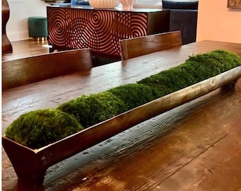 May include: A long, narrow, dark brown wooden dough bowl filled with vibrant green preserved moss. The bowl has a smooth, elongated rectangular shape with tapered ends and sits directly on a matching dark brown wooden dining table surface.
