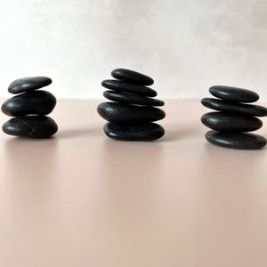 May include: Three stacks of smooth, black stones are arranged on a light pink surface. Each stack varies in height, with the tallest having six stones. The stones are oval-shaped and appear to be polished.