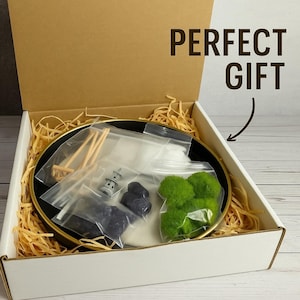 May include: An open box with a black and gold plate filled with a zen garden kit. The kit includes moss, rocks, a rake, a pagoda, and sand. The box is filled with brown packing paper. The text "PERFECT GIFT" is written on the box.