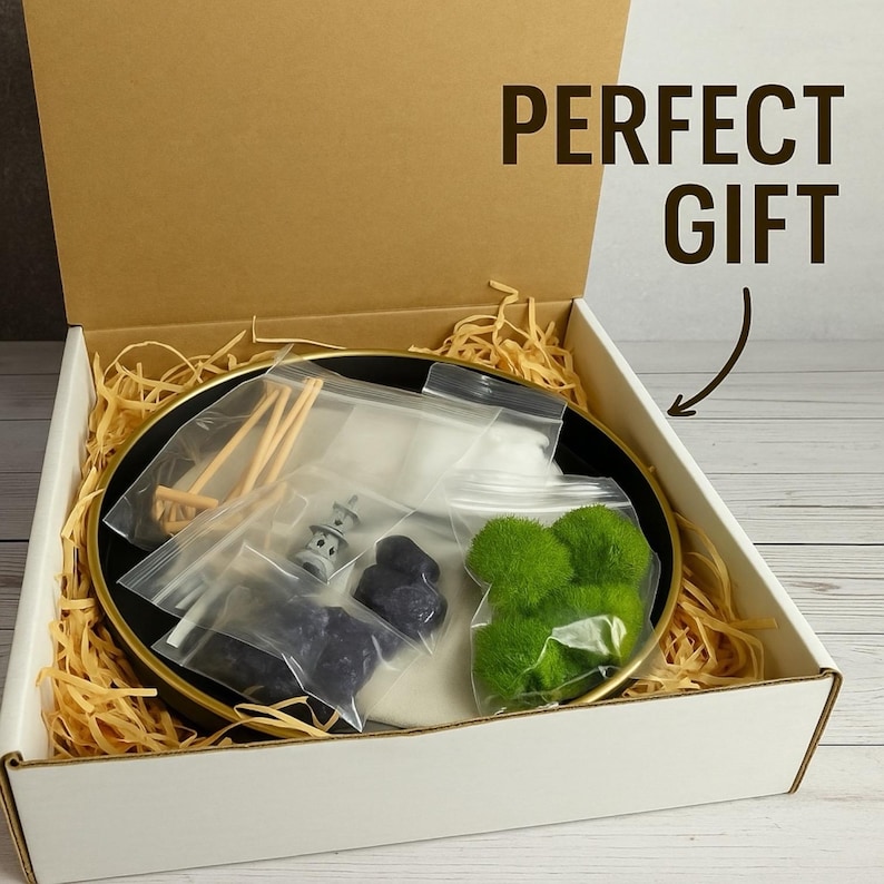 May include: An open box with a black and gold plate filled with a zen garden kit. The kit includes moss, rocks, a rake, a pagoda, and sand. The box is filled with brown packing paper. The text "PERFECT GIFT" is written on the box.