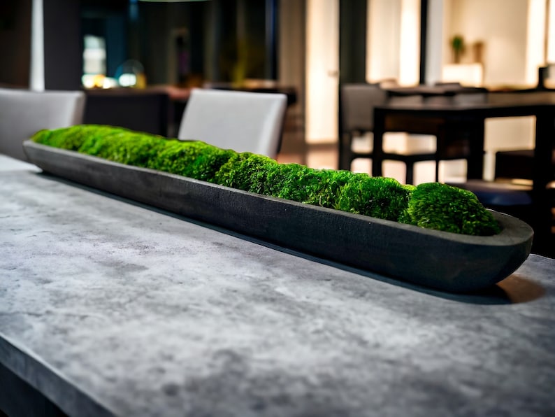 May include: A long, narrow, matte black rectangular trough planter filled with vibrant green preserved moss. The container has a smooth, solid construction and is positioned horizontally, showcasing a dense, textured arrangement of natural moss.