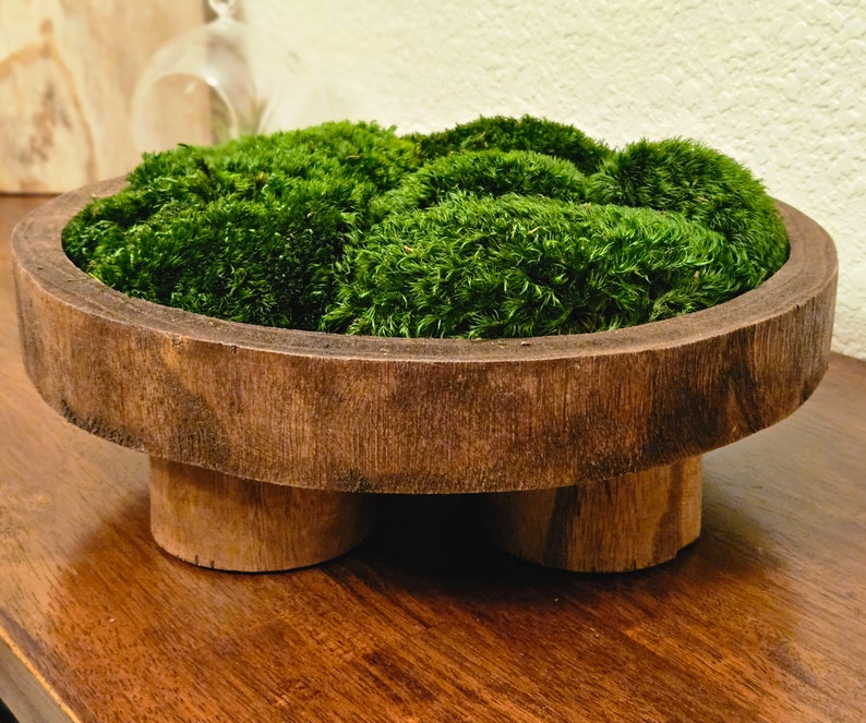 May include: A round, dark brown wooden bowl with a thick rim and three short, cylindrical wooden legs. The interior of the bowl is filled with a dense, vibrant green preserved moss that rises slightly above the top edge of the wooden container.