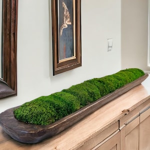 May include: A long, narrow, dark brown wooden dough bowl filled with vibrant green preserved moss. The bowl has a rustic, hand-carved appearance with visible wood grain and texture, creating a natural contrast against the dense, textured moss.