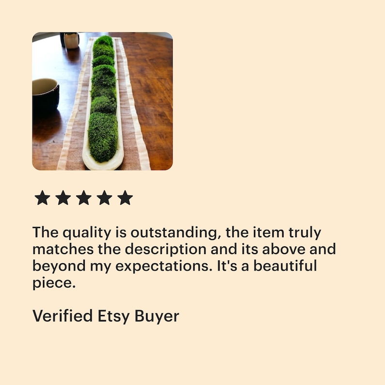 May include: A long, narrow, off-white ceramic trough-style bowl filled with a series of rounded, vibrant green moss clusters. The bowl is positioned lengthwise on a rectangular, light-colored fabric runner with subtle brown striped accents.