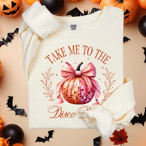 以下が含まれることがあります： オレンジとピンクの文字で「Take Me To The Disco Pumpkin」と書かれた白いスウェットシャツ。ピンクのリボンがディスコボールの南瓜の上に置かれています。