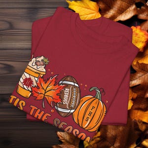 May include: Red maroon t-shirt with a graphic design of a pumpkin, football, maple leaf, and a coffee cup. The text on the shirt reads "Tis the season".